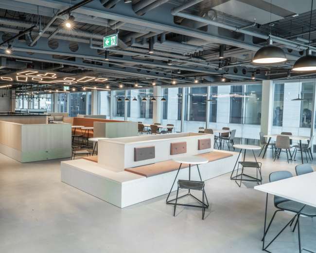The image shows a modern office space with an open layout featuring a café area, seating arrangements, and large windows providing natural light.