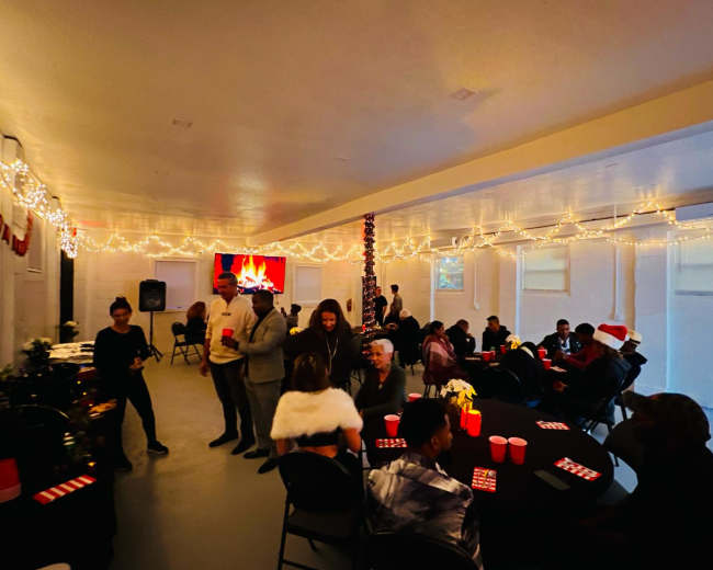 A decorated indoor venue filled with people socializing around tables, featuring festive lighting and a screen displaying a fireplace image.