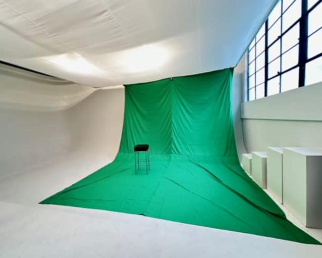 A green backdrop with a black stool is set up in a spacious studio with white walls and large windows.