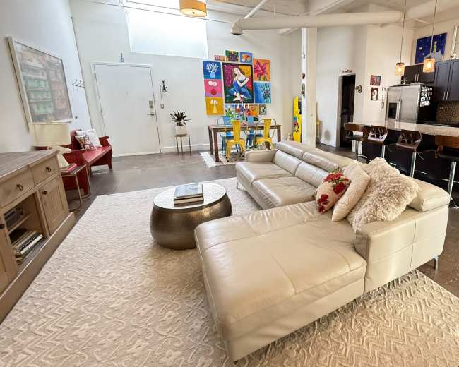 The image shows a modern living room with a light-colored sectional sofa, a round coffee table, a variety of colorful artwork on the wall, and a kitchen area in the background.