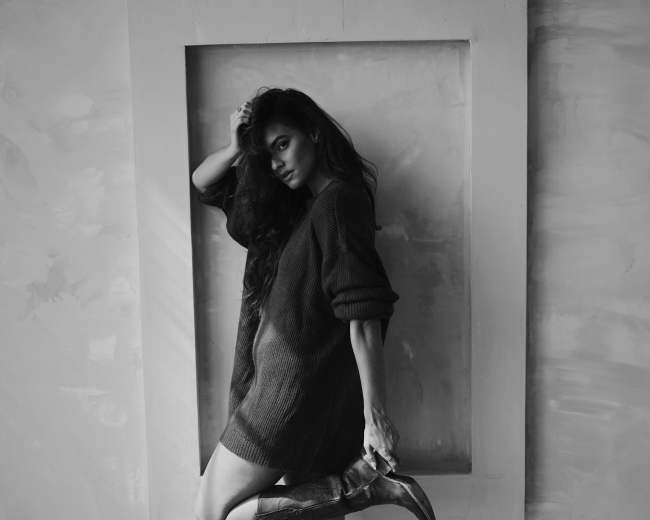 A woman poses in a dark sweater and cowboy boots against a textured wall framed by a white rectangular outline.
