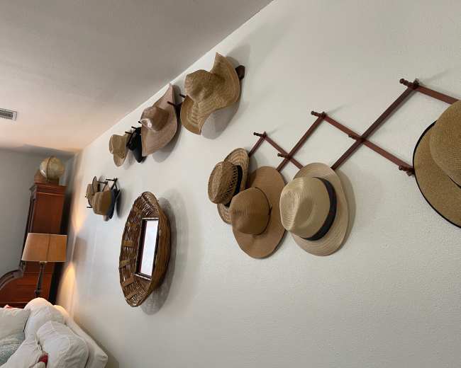 The image shows a wall with a variety of hats hanging on wooden hat racks, alongside a round wicker basket and a mirror.