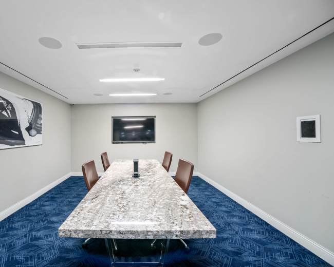 A modern conference room features a large granite table surrounded by four brown chairs, with a television mounted on the wall and abstract artwork displayed.