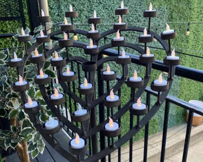 A heart-shaped metal stand holds multiple candles on a balcony surrounded by greenery.