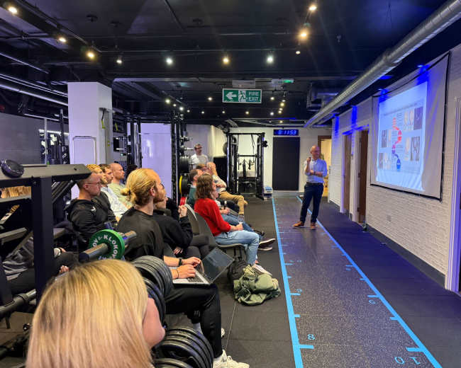 A speaker presents to an audience seated in a gym, with exercise equipment visible in the background and a presentation projected on a screen.