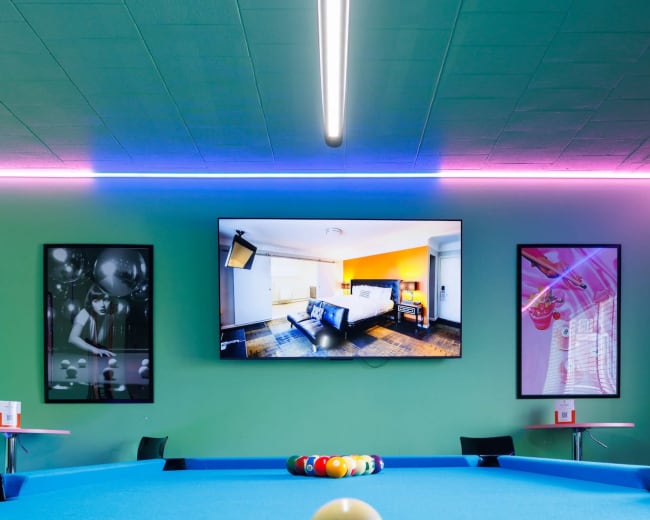 A pool table with a single cue ball sits in front of a colorful room featuring a large television and framed artwork on the walls.