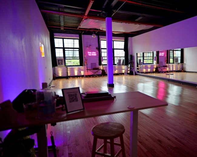 An empty dance studio with wooden floors, large mirrors, and purple lighting, featuring a small table and stool in the foreground.