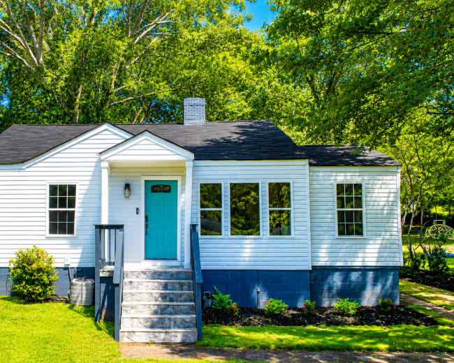 The image shows a white single-story house with a blue front door, surrounded by greenery and a well-maintained lawn.