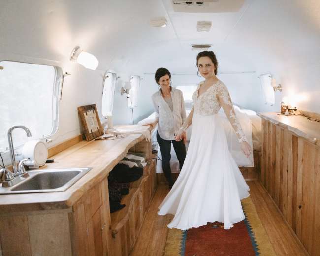 A bride in a white dress twirls in a rustic interior space while a woman stands nearby, smiling.