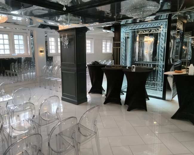 The image shows a modern event space with transparent chairs arranged in rows and two black cocktail tables near a mirrored wall.