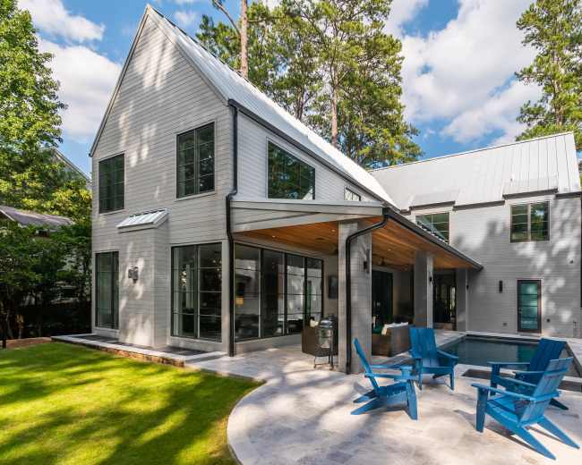 The image shows a contemporary house with a gray exterior, large windows, and a spacious patio area featuring blue Adirondack chairs beside a small pool.
