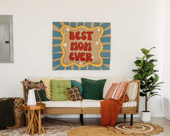 A cozy living room features a rattan couch adorned with colorful pillows, a decorative rug on a light floor, and a large wall art piece that reads "BEST MOM EVER."