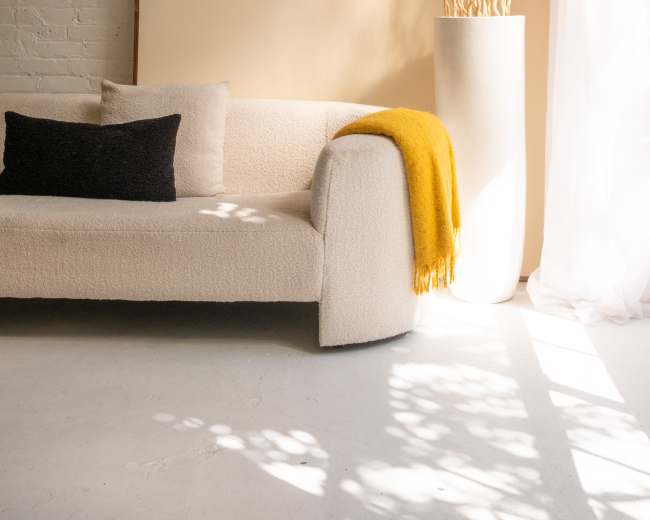 A modern, minimalist living space features a light-colored sofa with a black pillow, a yellow throw blanket, a tall white vase with decorative branches, and a large art piece against a textured wall.