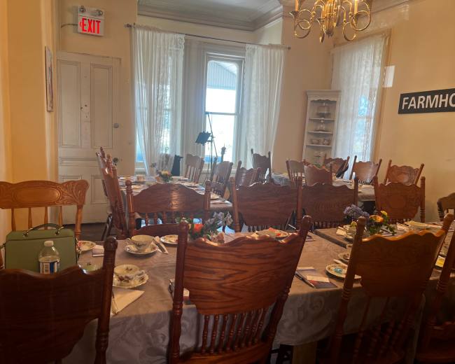 A dining room is set with a large table surrounded by wooden chairs, featuring floral centerpieces and table settings, with light streaming through the windows.