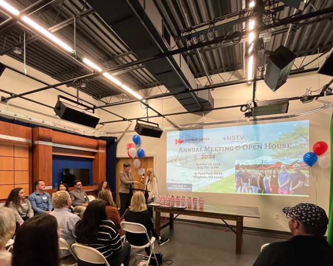 A group of people sits in a studio setting, attending an annual meeting and open house event, with a projector screen displaying event details and colorful balloons in the background.