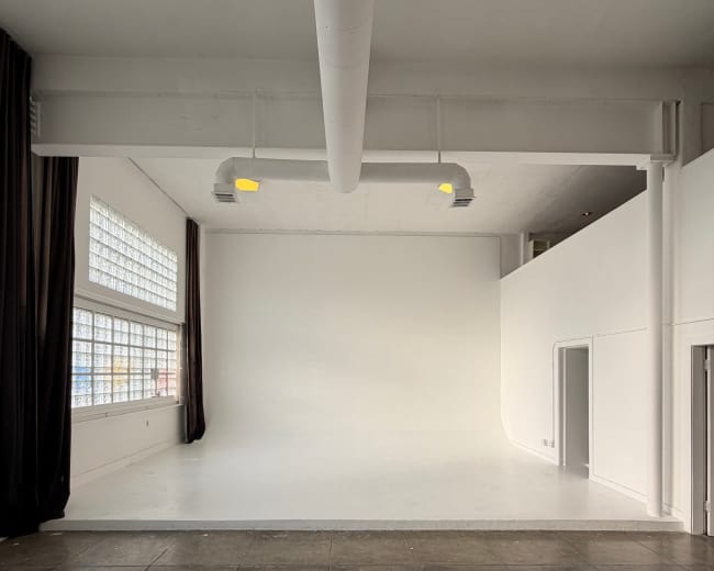 The image shows a spacious, empty room with white walls, a smooth white floor, large windows, and a tiled section on the floor.