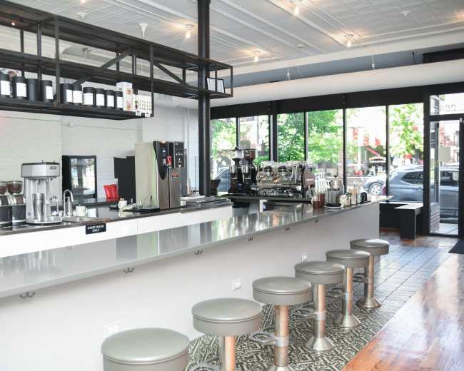 The image shows a modern café interior with a sleek counter, several bar stools, and coffee equipment lined up behind the counter.