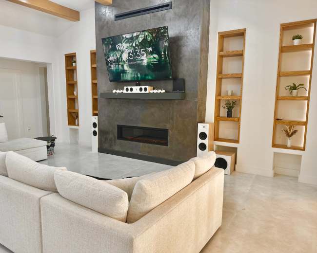 A modern living room features a large sectional sofa facing a mounted television above a sleek fireplace, with built-in shelves on either side displaying decorative plants.