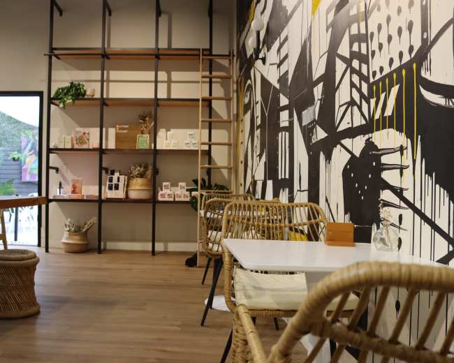 The image shows a modern café interior with a wall featuring abstract art, wooden shelves displaying products, and rattan furniture arranged around white tables.