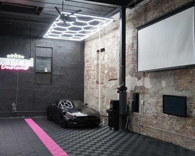 The image shows an industrial-style interior space with exposed brick walls, a black sports car parked on a checkered floor, and a neon sign that reads "Wrapped."