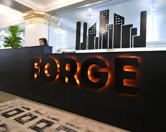 A receptionist stands behind a modern black front desk that features the illuminated word "FORGE" and a city skyline graphic above.