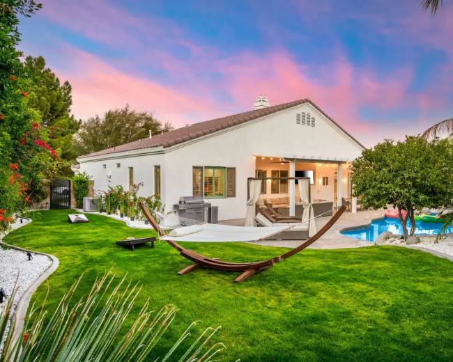 A white house with large windows is set in a landscaped yard featuring a hammock, swimming pool, and various plants against a colorful sunset sky.
