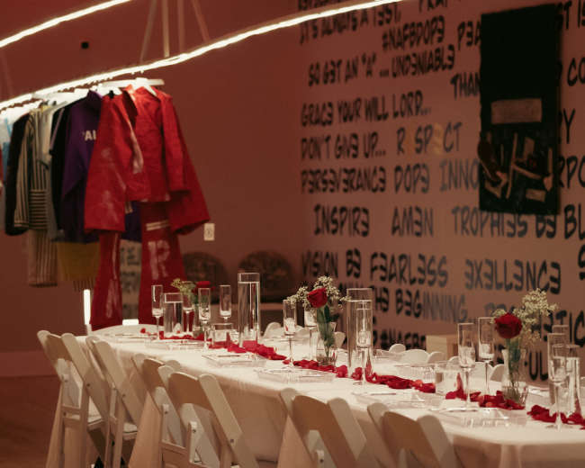 A long dining table set with glasses and rose petals is positioned under hanging clothing displays in a designed room.