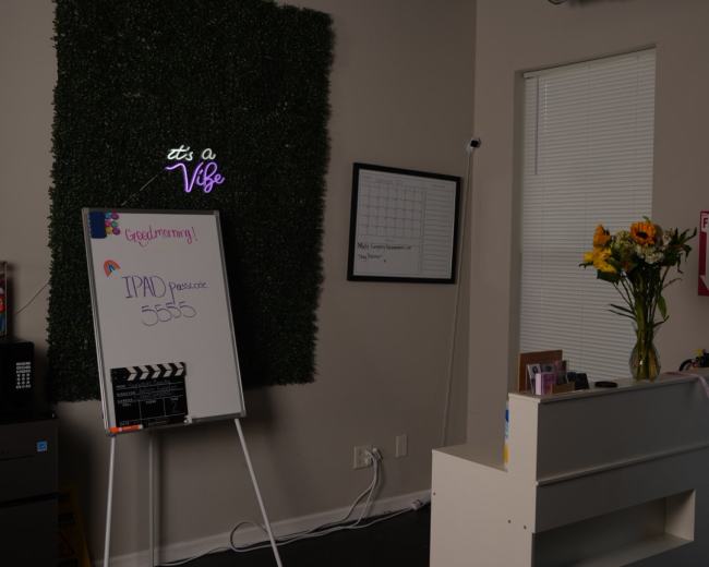 The image shows a room with a green wall decoration, a whiteboard displaying a message, a vase of flowers on a counter, and a small black filing cabinet.