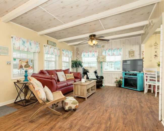 The image shows a living room with a red sofa, a light wooden coffee table, and a turquoise TV cabinet, surrounded by large windows with patterned curtains.