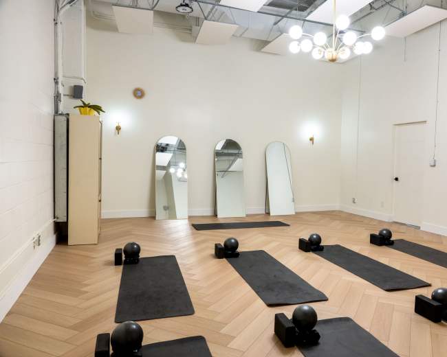 A bright, empty fitness studio features wooden flooring, exercise mats arranged on the ground, and mirrors hanging on the wall.