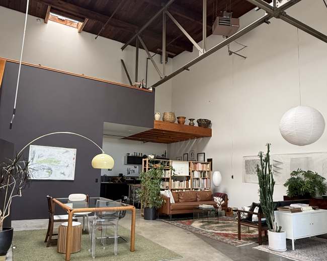 A spacious, modern interior features a dining area with a glass table and wooden chairs, a comfortable seating area with a sofa, and large plants, all under a high ceiling with exposed beams.