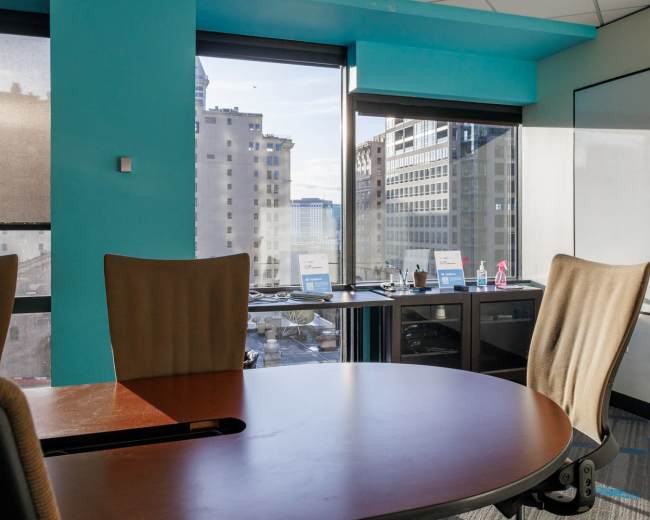 The image shows a conference room with a round wooden table and light-colored chairs, overlooking a cityscape through large windows.