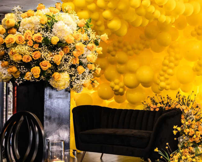 A yellow balloon installation surrounds a dark, velvet couch with floral arrangements in the foreground.