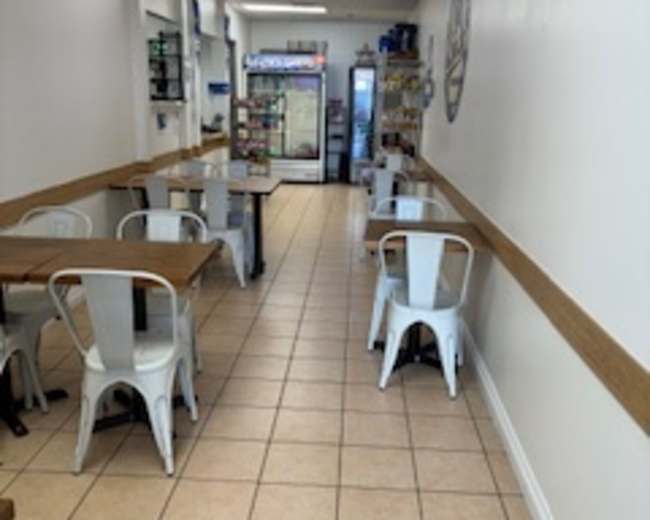 The image shows a narrow dining area with metal tables and chairs, lined with tiled flooring, and leading to a cooler at the far end.