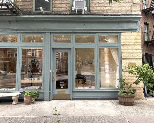 The image shows a storefront with large windows and a wooden door, framed by light blue trim, on a city street.