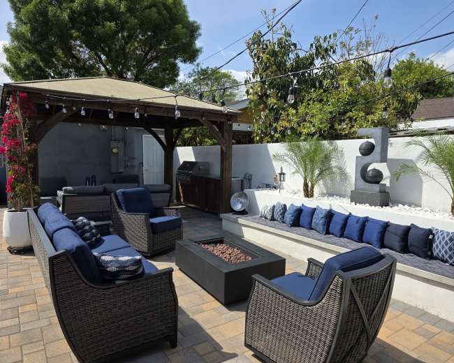 The image shows a stylish outdoor living area with a shaded seating arrangement, a barbecue grill, and decorative landscaping elements, including plants and a stone sculpture.