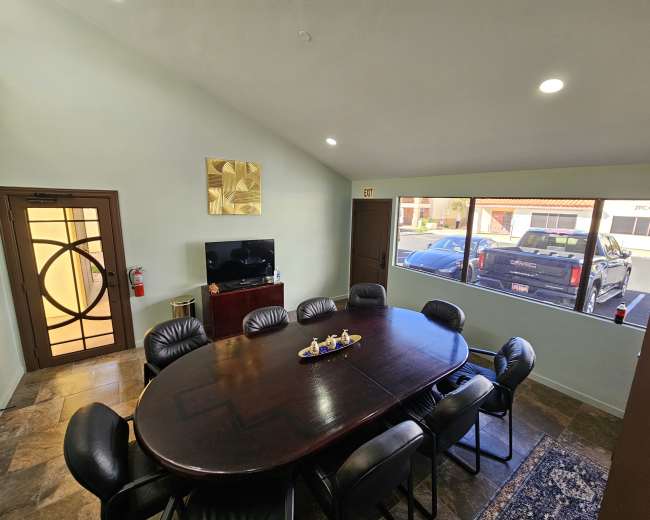 The image shows a conference room with a large oval table surrounded by black leather chairs, a television mounted on the wall, and a view of parked vehicles outside through a large window.