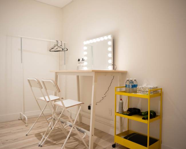 The image shows a minimalist dressing room with a mirror surrounded by lights, a counter with two folding chairs, and a yellow cart holding water bottles and other items.
