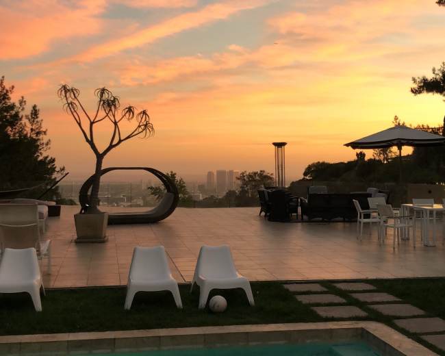 The scene shows a swimming pool surrounded by white chairs and a sunset sky with colorful clouds over a cityscape.