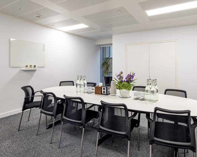 A conference room features a white oval table surrounded by black chairs, with glass water bottles and a flower arrangement in the center.