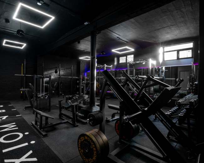 The image shows a dimly lit gym equipped with various weightlifting machines and free weights, featuring modern lighting.