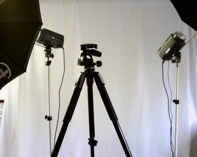 The image shows a photography setup with a tripod in the center, flanked by two studio lights and a backdrop.