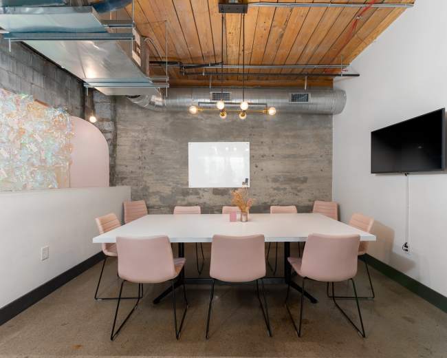 A modern conference room features a long white table surrounded by pink chairs, with exposed wooden beams and a large screen on the wall.
