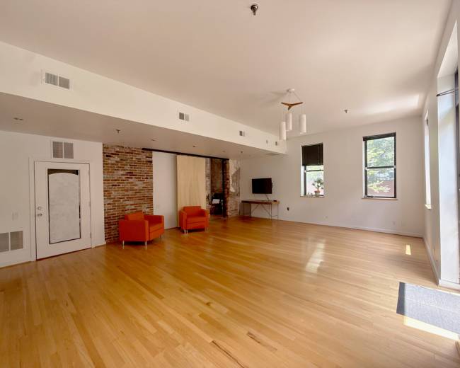 The image shows a spacious living area with hardwood floors, two orange armchairs, a television, and exposed brick walls.