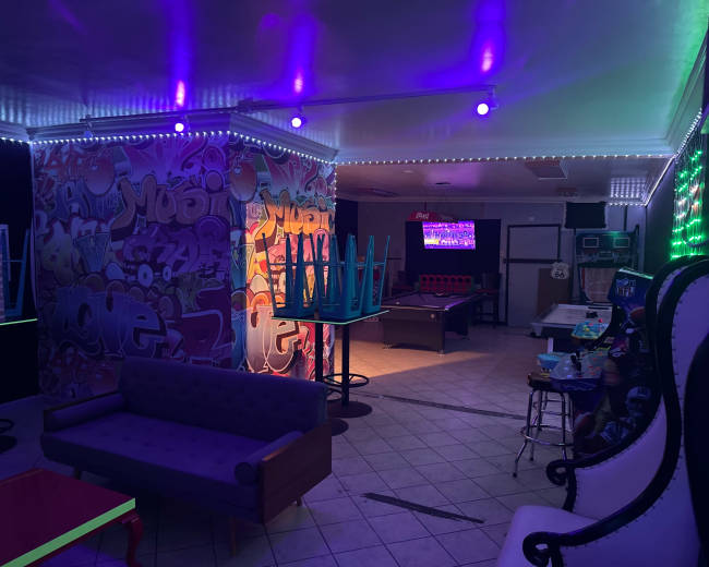 The image shows a dimly lit lounge area featuring colorful graffiti on the walls, stacked chairs, a pool table, and neon lighting accents.