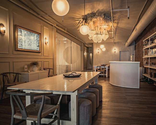 The image showcases a modern interior space featuring a long dining table, various seating options, a bar area, and decorative lighting against exposed brick walls.