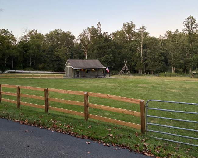 A wooden cabin is located in a grassy field surrounded by a wooden fence, with a group of trees in the background and a tepee structure nearby.