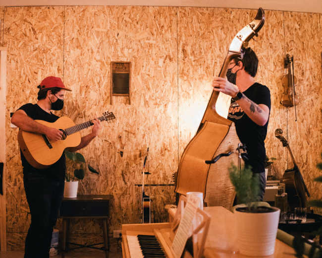 Two musicians play instruments—a guitarist holding an acoustic guitar and a bassist—with wooden walls and various musical instruments in the background.