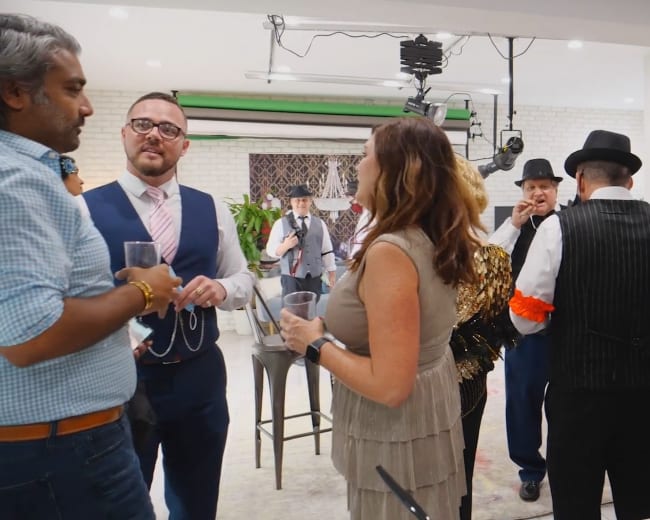 A group of people in costumes socializes at a festive indoor gathering, with a table set with food and drinks in the foreground.