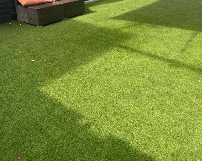The image shows a backyard space with artificial grass, a black brick wall, a small couch with orange cushions, and a partially shaded area from a nearby structure.